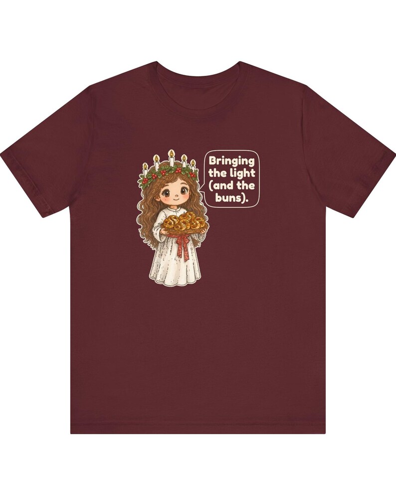 May include: A maroon t-shirt featuring a graphic of a character wearing a white robe and a candle crown, holding a tray of buns. A speech bubble next to the character contains the text: Bringing the light (and the buns).