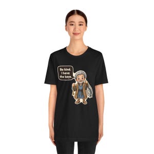 May include: A black t-shirt featuring a cartoon illustration of a gray-haired person in a blue tunic and brown robe holding a large key and a net. A speech bubble above the character contains the text: Be kind. I have the keys.