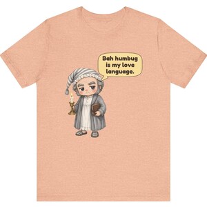 Puede incluir: Una camiseta color melocot&oacute;n con una ilustraci&oacute;n de dibujos animados de un personaje con gorro de dormir y bata que sostiene una vela y un libro. Un bocadillo sobre el personaje contiene el texto: "Bah humbug is my love language."