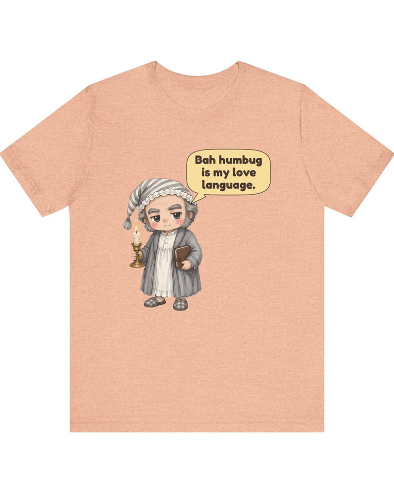 Puede incluir: Una camiseta color melocot&oacute;n con una ilustraci&oacute;n de dibujos animados de un personaje con gorro de dormir y bata que sostiene una vela y un libro. Un bocadillo sobre el personaje contiene el texto: "Bah humbug is my love language."