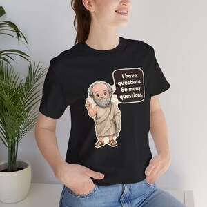May include: A black short-sleeved t-shirt featuring a cartoon illustration of a bearded man in a beige toga pointing upward. A speech bubble next to him contains the text, "I have questions. So many questions." The shirt has a crew neckline.