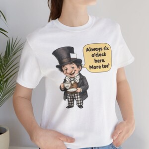 May include: A white t-shirt featuring a graphic of a character wearing a top hat, a polka dot bow tie, a dark jacket, and checkered pants, holding a teacup. A yellow speech bubble contains the text: Always six o'clock here. More tea?