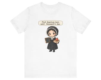 May include: A white short-sleeved t-shirt featuring a cartoon illustration of a figure in a black dress and bonnet holding a chalkboard and a book with an apple on top. A speech bubble above the figure contains the text: First American-born saint. Homework's due.