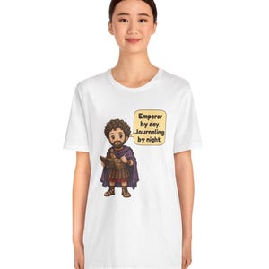 K&ouml;nnte beinhalten: Ein wei&szlig;es Kurzarm-T-Shirt mit einer Cartoon-Illustration eines b&auml;rtigen Mannes in r&ouml;mischer R&uuml;stung und violettem Umhang, der ein Tagebuch und einen Stift h&auml;lt. Eine Sprechblase enth&auml;lt den Text: Emperor by day. Journaling by night.