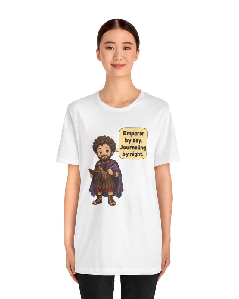 K&ouml;nnte beinhalten: Ein wei&szlig;es Kurzarm-T-Shirt mit einer Cartoon-Illustration eines b&auml;rtigen Mannes in r&ouml;mischer R&uuml;stung und violettem Umhang, der ein Tagebuch und einen Stift h&auml;lt. Eine Sprechblase enth&auml;lt den Text: Emperor by day. Journaling by night.