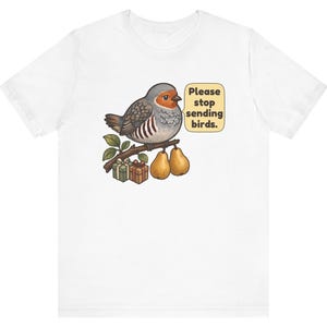 K&ouml;nnte beinhalten: Ein wei&szlig;es T-Shirt mit einer Grafik eines grauen und orangefarbenen Vogels, der auf einem Ast mit zwei Birnen und zwei kleinen Geschenkkartons sitzt. Eine Sprechblase neben dem Vogel enth&auml;lt den Text, Please stop sending birds.