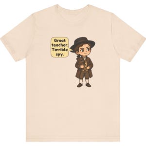 May include: A light beige t-shirt featuring a cartoon illustration of a person in 18th-century attire, including a brown coat, vest, and hat. A speech bubble next to the character contains the text: Great teacher. Terrible spy.