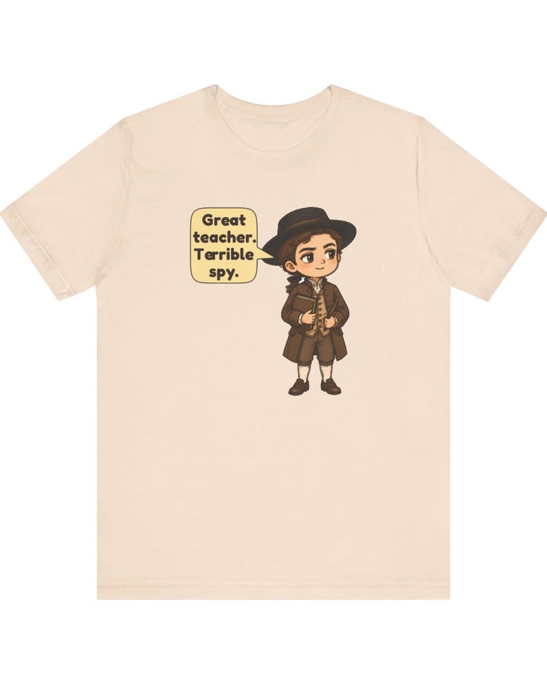 May include: A light beige t-shirt featuring a cartoon illustration of a person in 18th-century attire, including a brown coat, vest, and hat. A speech bubble next to the character contains the text: Great teacher. Terrible spy.