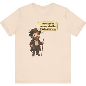 May include: A light beige t-shirt featuring a cartoon illustration of a bearded traveler in a brown hat and coat, holding a walking stick. A speech bubble above the character contains the text: I walked a thousand miles. Pack a lunch.