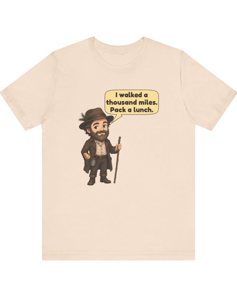 May include: A light beige t-shirt featuring a cartoon illustration of a bearded traveler in a brown hat and coat, holding a walking stick. A speech bubble above the character contains the text: I walked a thousand miles. Pack a lunch.
