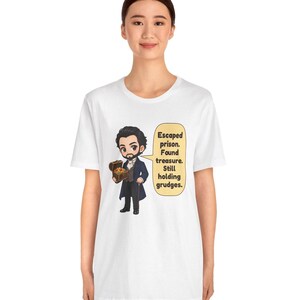 May include: A white t-shirt featuring a cartoon illustration of a bearded man in a dark coat holding a treasure chest. A speech bubble next to him contains the text: Escaped prison. Found treasure. Still holding grudges.
