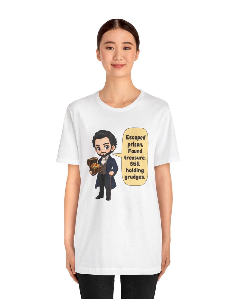 May include: A white t-shirt featuring a cartoon illustration of a bearded man in a dark coat holding a treasure chest. A speech bubble next to him contains the text: Escaped prison. Found treasure. Still holding grudges.