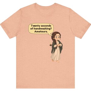 Op de afbeelding: Een perzikkleurig gem&ecirc;leerd T-shirt met een cartoonillustratie van een persoon met een lange bruine vlecht, gekleed in een cr&egrave;mekleurige jurk en donkere sjaal, die een kaars vasthoudt. Een tekstballon bevat de tekst: Twenty seconds of handwashing? Amateurs.