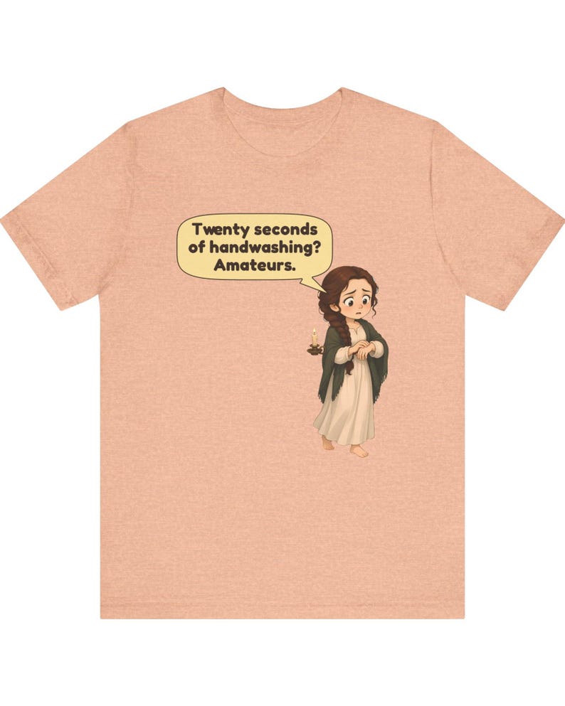 Op de afbeelding: Een perzikkleurig gem&ecirc;leerd T-shirt met een cartoonillustratie van een persoon met een lange bruine vlecht, gekleed in een cr&egrave;mekleurige jurk en donkere sjaal, die een kaars vasthoudt. Een tekstballon bevat de tekst: Twenty seconds of handwashing? Amateurs.