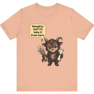 Puede incluir: Una camiseta de color melocot&oacute;n con un gr&aacute;fico de una peque&ntilde;a criatura marr&oacute;n oscuro con cuernos que sostiene un manojo de palos y un pergamino. Un bocadillo amarillo sobre la criatura contiene el texto: Naughty list? I'll take it from here.
