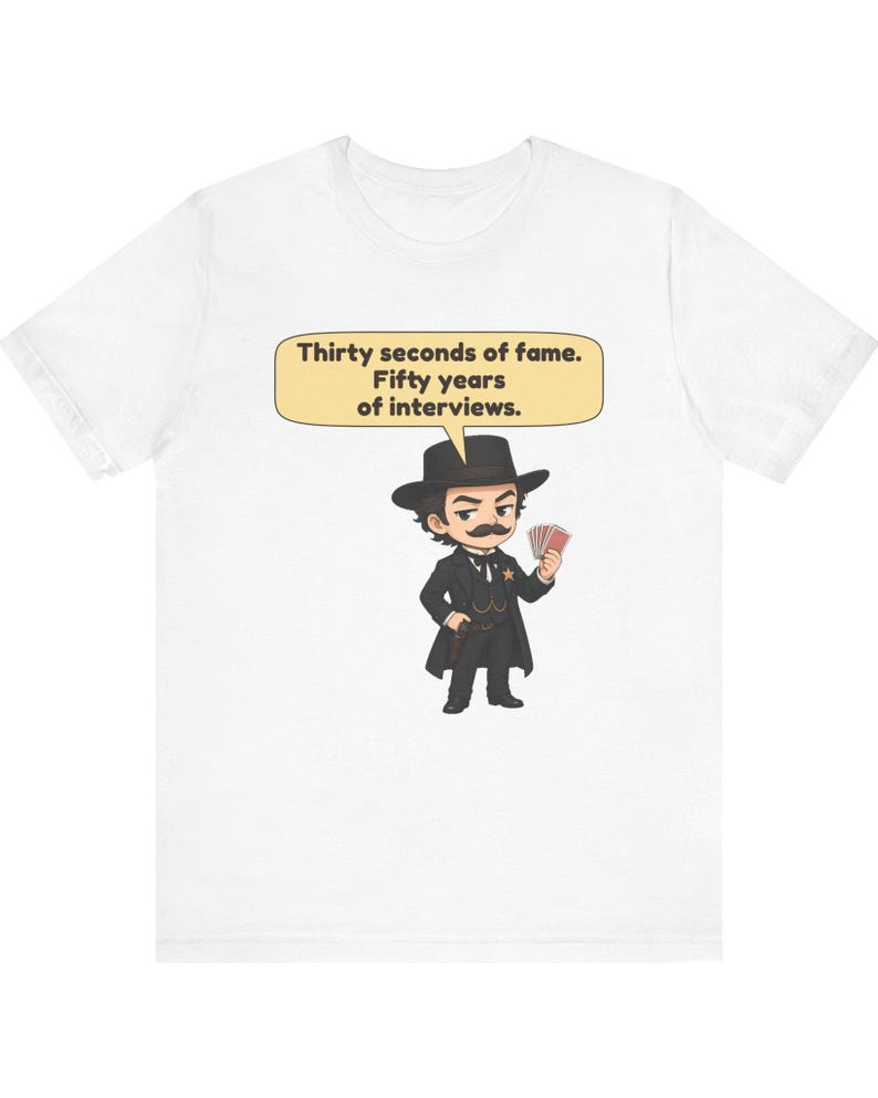 Pu&ograve; includere: Una t-shirt bianca con un'illustrazione a cartoni animati di un uomo con i baffi che indossa un cappello e un cappotto neri, mentre tiene in mano delle carte da gioco. Un fumetto contiene il testo: Thirty seconds of fame. Fifty years of interviews.