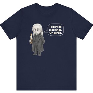 May include: A navy blue short-sleeve t-shirt featuring a graphic of a pale, long-haired character in a dark coat holding a lit candle. A speech bubble next to the character contains the text: I don't do mornings. Or garlic.