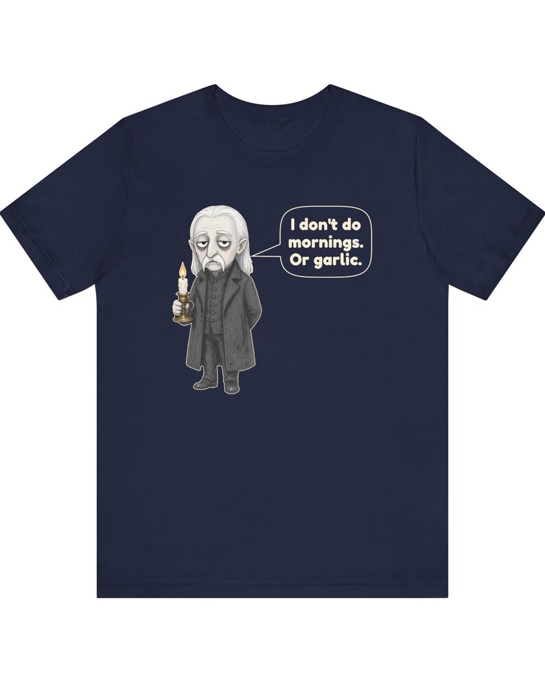 May include: A navy blue short-sleeve t-shirt featuring a graphic of a pale, long-haired character in a dark coat holding a lit candle. A speech bubble next to the character contains the text: I don't do mornings. Or garlic.