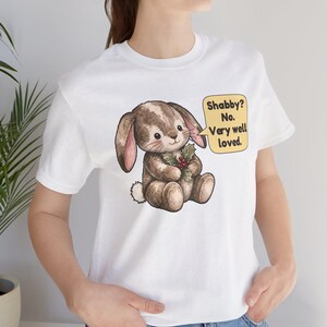 May include: A white t-shirt featuring a brown plush bunny illustration holding a sprig of holly. A yellow speech bubble next to the bunny contains the text: Shabby? No. Very well loved. The bunny has stitched details and a small white tail.