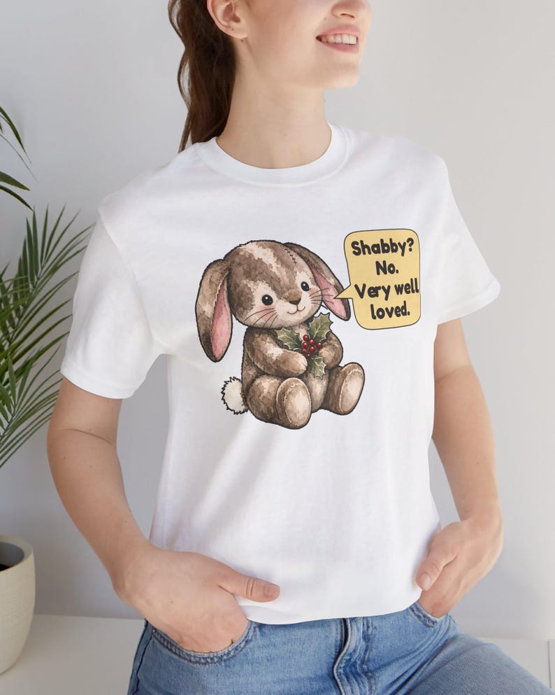 May include: A white t-shirt featuring a brown plush bunny illustration holding a sprig of holly. A yellow speech bubble next to the bunny contains the text: Shabby? No. Very well loved. The bunny has stitched details and a small white tail.