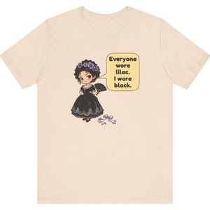 May include: A beige short-sleeve t-shirt featuring a cartoon illustration of a character in a black dress with a lilac flower crown and fan. A speech bubble contains the text: Everyone wore lilac. I wore black. A small lilac ribbon lies below.