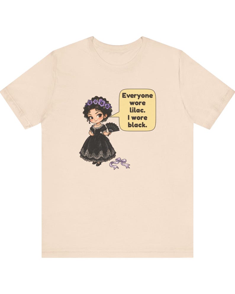 May include: A beige short-sleeve t-shirt featuring a cartoon illustration of a character in a black dress with a lilac flower crown and fan. A speech bubble contains the text: Everyone wore lilac. I wore black. A small lilac ribbon lies below.