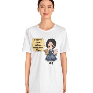 May include: A white t-shirt featuring a cartoon illustration of a person in a blue Victorian-style dress holding punch cards. A speech bubble next to the character contains the text: I wrote code before computers. Flex.