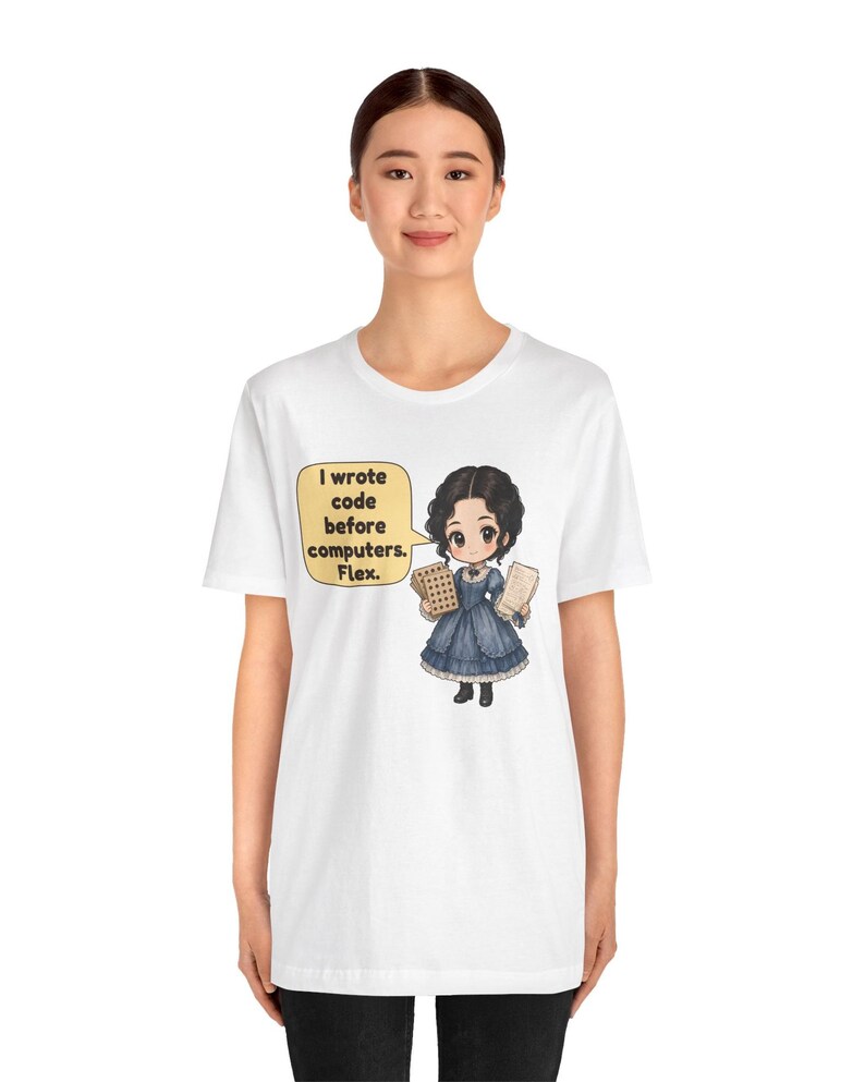 May include: A white t-shirt featuring a cartoon illustration of a person in a blue Victorian-style dress holding punch cards. A speech bubble next to the character contains the text: I wrote code before computers. Flex.