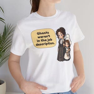 Op de afbeelding: Een wit t-shirt met korte mouwen en een afbeelding van een vrouw en een kind met een kaars, vergezeld door een klein spookje. Een gele tekstballon bevat de zwarte tekst: Ghosts weren't in the job description.