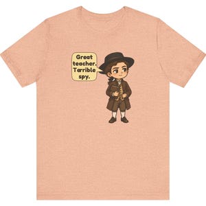 May include: A peach-colored t-shirt featuring a cartoon illustration of a person in 18th-century attire, including a brown coat, hat, and breeches. A speech bubble next to the character contains the text: Great teacher. Terrible spy.