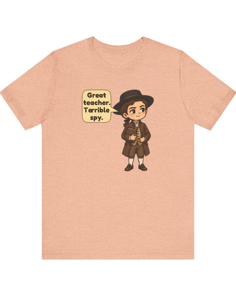 May include: A peach-colored t-shirt featuring a cartoon illustration of a person in 18th-century attire, including a brown coat, hat, and breeches. A speech bubble next to the character contains the text: Great teacher. Terrible spy.