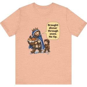 May include: A peach-colored t-shirt featuring a cartoon illustration of a person wearing a crown and blue cloak carrying a basket of food, accompanied by a child. A speech bubble contains the text: Brought dinner through snow. No tip.