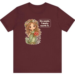 May include: A maroon short-sleeve t-shirt featuring a graphic of a cartoon character with long brown hair, a flower crown, and a green dress holding a pomegranate. A speech bubble next to the character contains the text: Six seeds. Totally worth it.