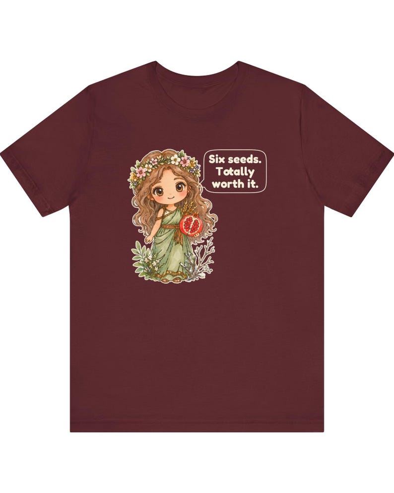 May include: A maroon short-sleeve t-shirt featuring a graphic of a cartoon character with long brown hair, a flower crown, and a green dress holding a pomegranate. A speech bubble next to the character contains the text: Six seeds. Totally worth it.