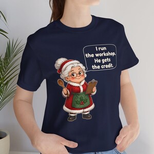 K&ouml;nnte beinhalten: Ein marineblaues T-Shirt mit einer Grafik einer &auml;lteren Frau mit wei&szlig;em Haar, Brille und einem roten Kleid mit gr&uuml;ner Sch&uuml;rze. Sie h&auml;lt einen Holzl&ouml;ffel und ein Klemmbrett. Eine Sprechblase enth&auml;lt den Text: I run the workshop. He gets the credit.