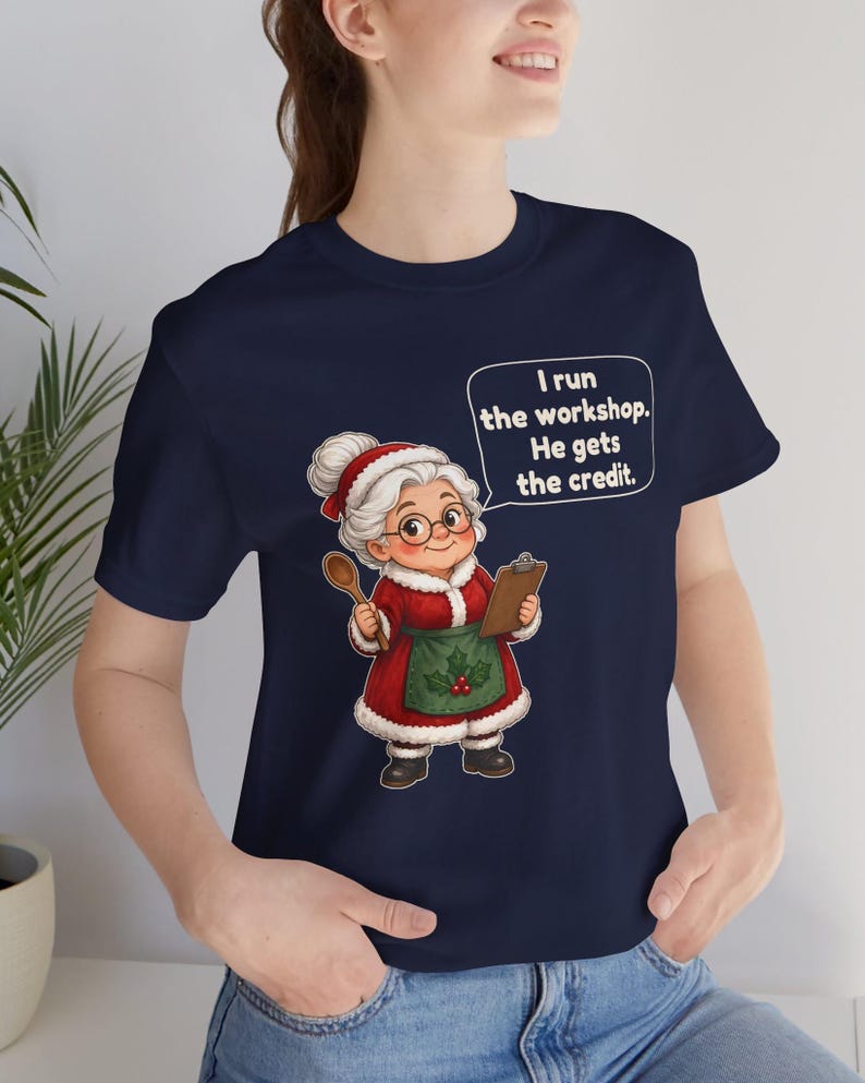 K&ouml;nnte beinhalten: Ein marineblaues T-Shirt mit einer Grafik einer &auml;lteren Frau mit wei&szlig;em Haar, Brille und einem roten Kleid mit gr&uuml;ner Sch&uuml;rze. Sie h&auml;lt einen Holzl&ouml;ffel und ein Klemmbrett. Eine Sprechblase enth&auml;lt den Text: I run the workshop. He gets the credit.