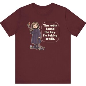 May include: A maroon t-shirt featuring a graphic of a person in a dark coat holding a large key with a robin perched on it. The text in a speech bubble reads, "The robin found the key. I'm taking credit." Small purple flowers grow near the person's boots.