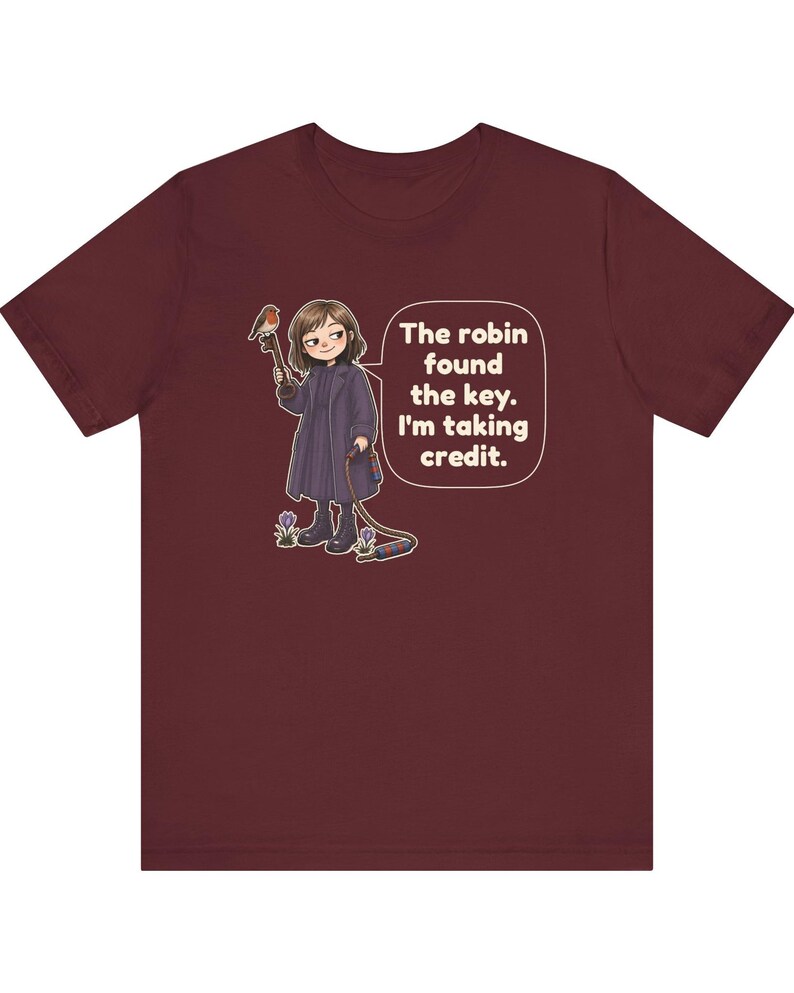 May include: A maroon t-shirt featuring a graphic of a person in a dark coat holding a large key with a robin perched on it. The text in a speech bubble reads, "The robin found the key. I'm taking credit." Small purple flowers grow near the person's boots.