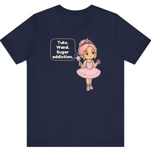 May include: A navy blue t-shirt featuring a graphic of a cartoon ballerina with pink hair, a tiara, and a pink tutu. The character holds a star-tipped wand and a cupcake. A speech bubble contains the text: Tutu. Wand. Sugar addiction.