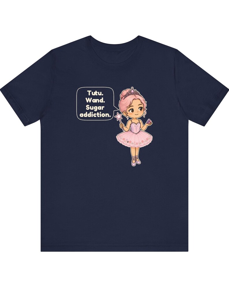 May include: A navy blue t-shirt featuring a graphic of a cartoon ballerina with pink hair, a tiara, and a pink tutu. The character holds a star-tipped wand and a cupcake. A speech bubble contains the text: Tutu. Wand. Sugar addiction.
