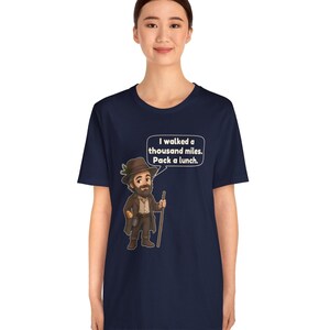 May include: A navy blue t-shirt featuring a cartoon illustration of a bearded traveler wearing a brown hat and coat, holding a walking stick. A speech bubble above the character contains the text: I walked a thousand miles. Pack a lunch.