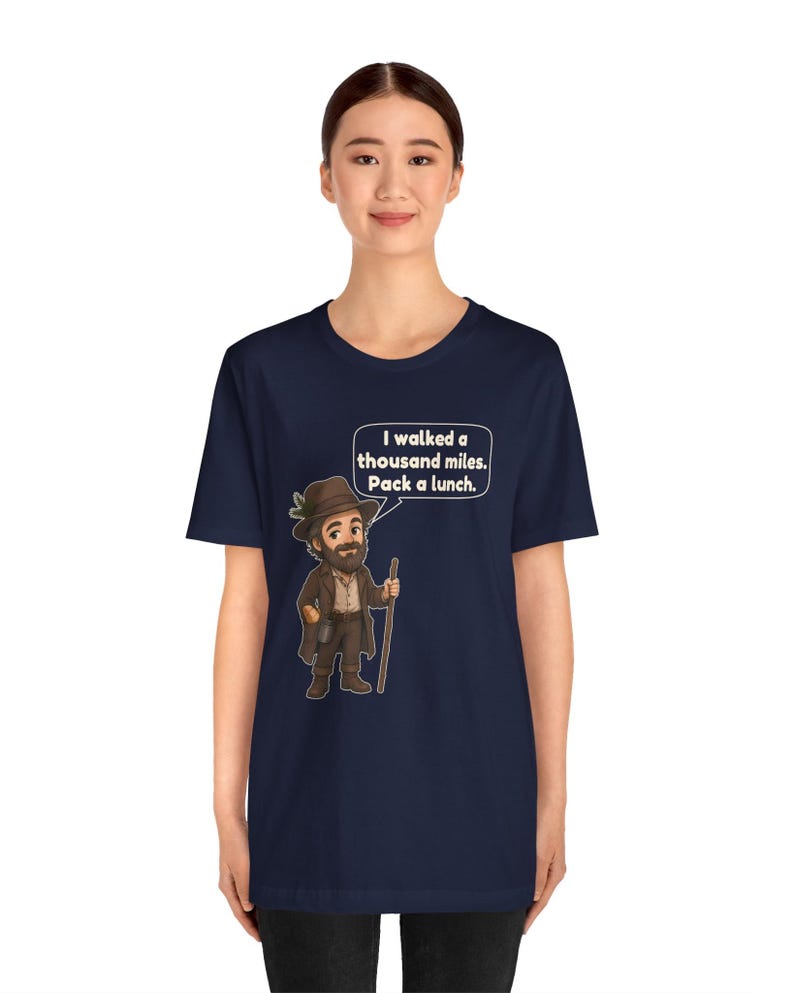 May include: A navy blue t-shirt featuring a cartoon illustration of a bearded traveler wearing a brown hat and coat, holding a walking stick. A speech bubble above the character contains the text: I walked a thousand miles. Pack a lunch.