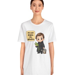 May include: A white t-shirt featuring a cartoon illustration of a person in a black robe holding green peas and a watering can. A speech bubble next to the character contains the text: It's not drama. It's genetics.