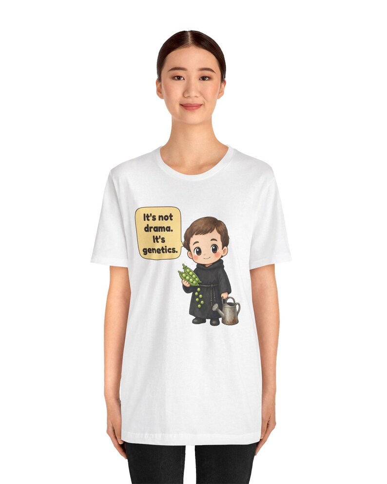 May include: A white t-shirt featuring a cartoon illustration of a person in a black robe holding green peas and a watering can. A speech bubble next to the character contains the text: It's not drama. It's genetics.