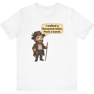 May include: A white t-shirt featuring a cartoon illustration of a bearded traveler wearing a brown hat with a green sprig, a brown coat, and boots. The character holds a wooden walking stick and a speech bubble that reads: I walked a thousand miles. Pack a lunch.