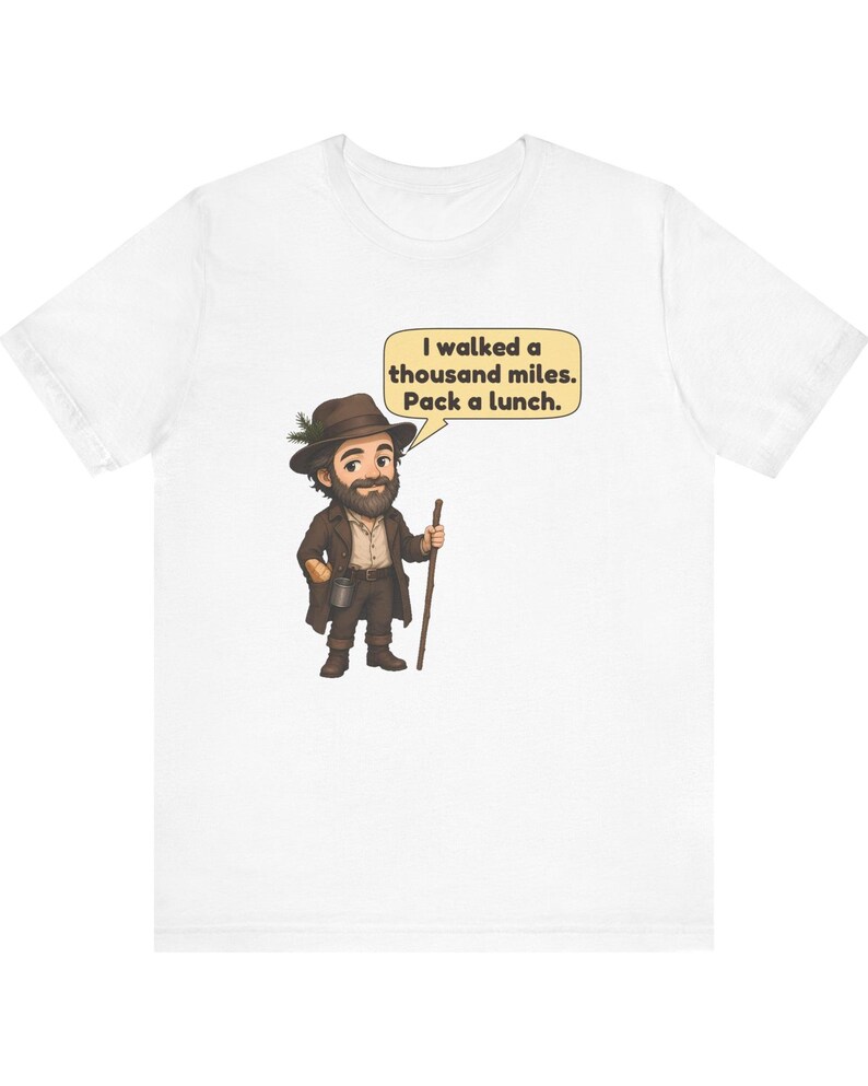 May include: A white t-shirt featuring a cartoon illustration of a bearded traveler wearing a brown hat with a green sprig, a brown coat, and boots. The character holds a wooden walking stick and a speech bubble that reads: I walked a thousand miles. Pack a lunch.