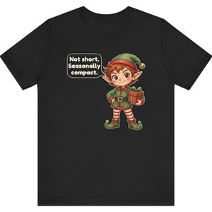 May include: A black t-shirt featuring a cartoon elf wearing a green outfit with red and white striped leggings, holding a red gift box with a green ribbon. A speech bubble above the elf contains the text: Not short. Seasonally compact.