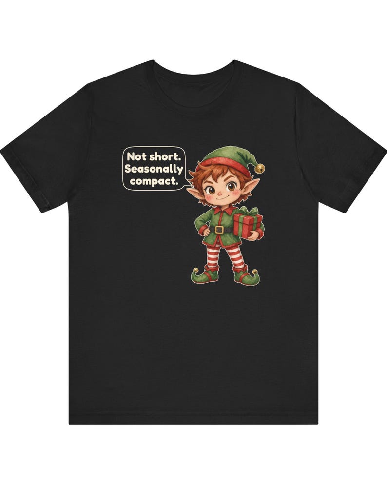 May include: A black t-shirt featuring a cartoon elf wearing a green outfit with red and white striped leggings, holding a red gift box with a green ribbon. A speech bubble above the elf contains the text: Not short. Seasonally compact.