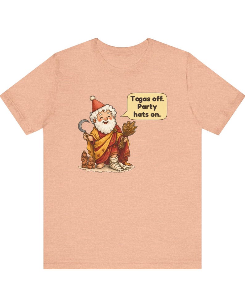May include: A peach-colored t-shirt featuring a cartoon illustration of a bearded figure in a red and yellow robe, holding a sickle and wheat, wearing a party hat, with a speech bubble that reads: Togas off. Party hats on.