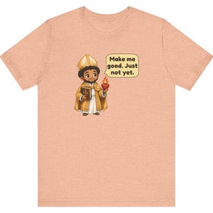 May include: A peach-colored t-shirt featuring a cartoon illustration of a figure in gold religious vestments holding a book and a flaming heart. A speech bubble next to the figure contains the text: Make me good. Just not yet.